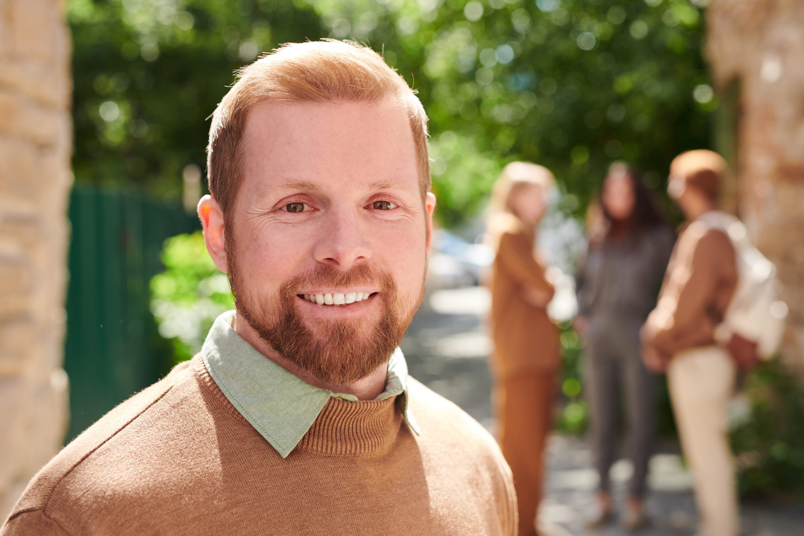 Portrait of smiling handsome young bearded college teacher in sweater standing against campus